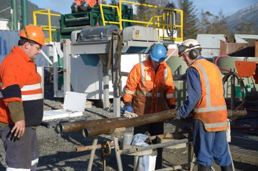 Trzech pracowników w odblaskowych ubraniach i kaskach pracuje przy rurach w terenie, w tle widoczna maszyna wiertnicza i drzewa.