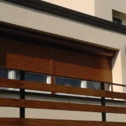 Zamknięte, brązowe rolety zewnętrzne na oknie z balkonem, widoczna antena satelitarna na ścianie budynku.