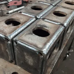 Spawane, metalowe bloki z otworem, ułożone na stole spawalniczym, widoczne narzędzia w tle.