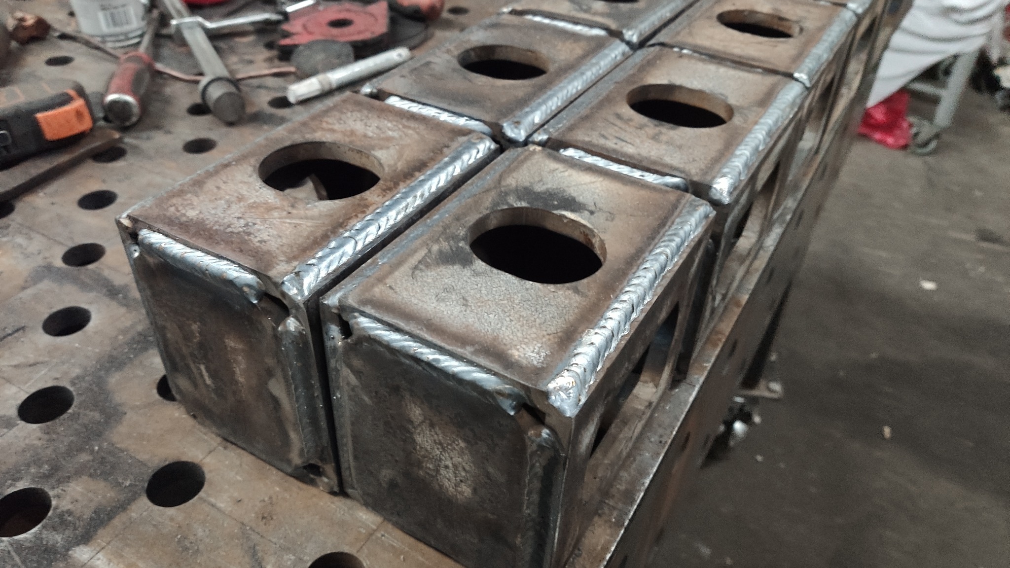Spawane, metalowe bloki z otworem, ułożone na stole spawalniczym, widoczne narzędzia w tle.