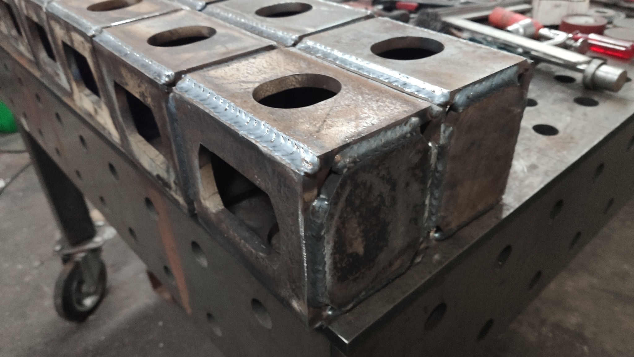 Spawane metalowe kostki z otworami, ułożone w rzędzie na perforowanym stole spawalniczym z kółkami. Widoczne ślady spawania i obróbki metalu.