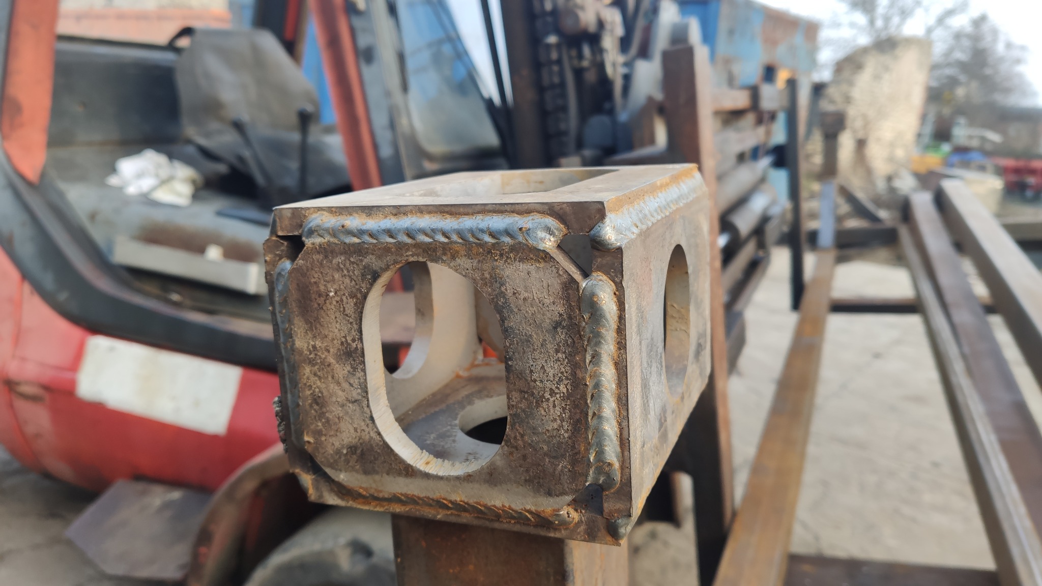 Spawana, metalowa konstrukcja z otworami, prawdopodobnie element wózka widłowego, widoczny w tle wózek widłowy.