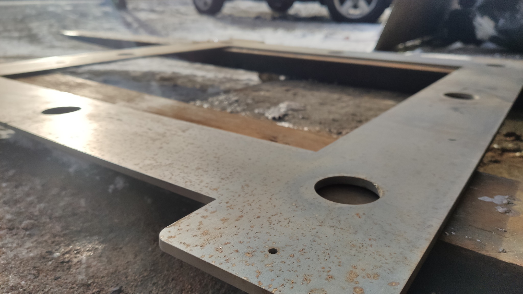 Metalowa rama z otworami, lekko zardzewiała, leżąca na ziemi, w tle fragment samochodu.