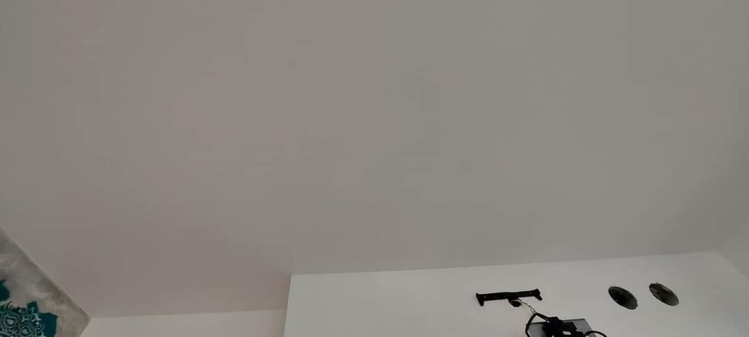 Biały sufit po malowaniu z widocznymi otworami na oświetlenie i fragmentem ściany z dekoracyjną tapetą w lewym dolnym rogu.