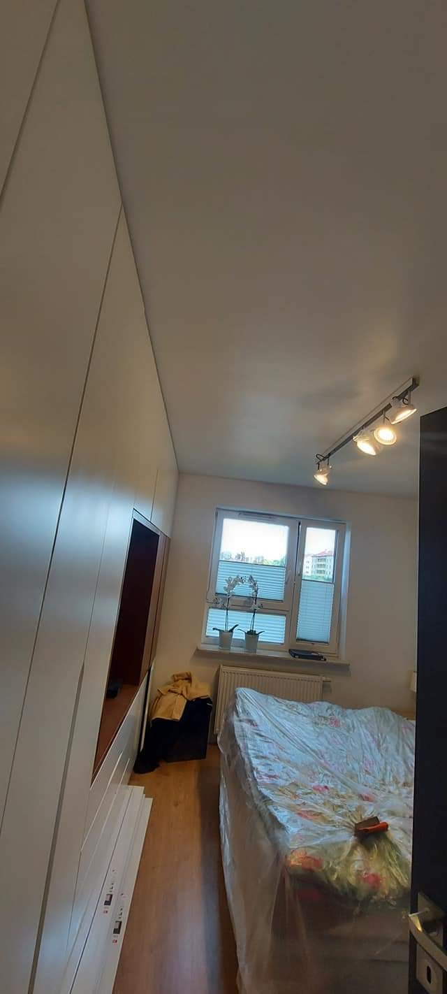 Nowoczesna szafa wnękowa z białymi frontami i otwartą półką w pokoju z zabezpieczonym folią łóżkiem podczas prac wykończeniowych, widoczne listwy przypodłogowe i oświetlenie sufitowe.