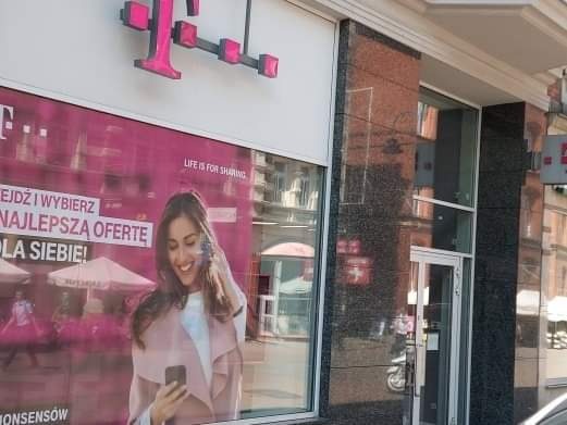 Fasada budynku z dużym oknem wystawowym, na którym wisi plakat reklamowy z uśmiechniętą kobietą trzymającą telefon, logo operatora telekomunikacyjnego na górze, marmurowa okładzina ściany obok.