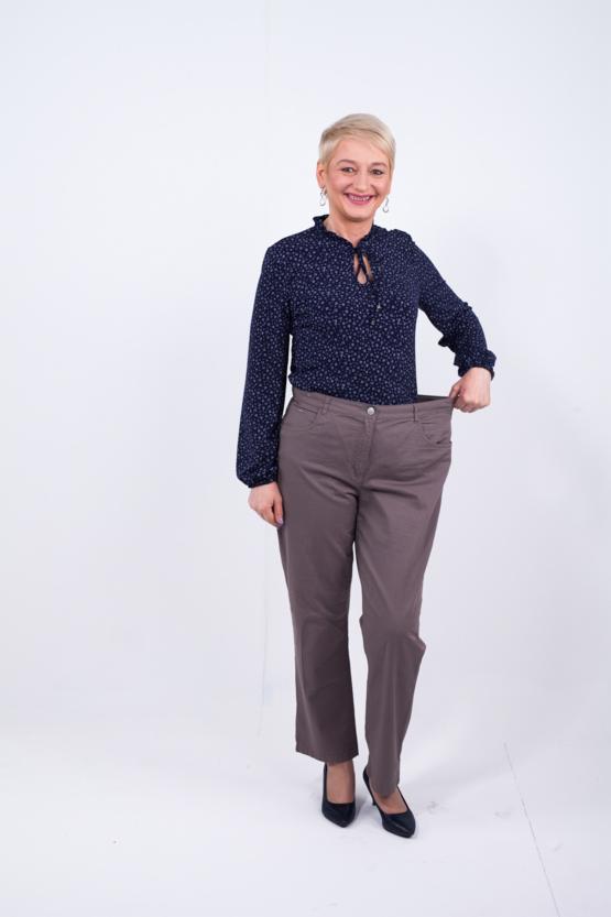 Uśmiechnięta kobieta prezentuje spodnie, które stały się za duże po utracie wagi, trzymając je odsunięte od ciała, ukazując luźny krój w pasie.