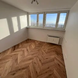 Pokój po remoncie z parkietem ułożonym we wzór jodełki, białe ściany, grzejnik pod oknem z widokiem na miasto.