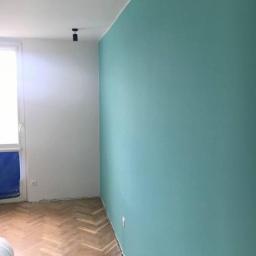 BK Wilbud Sp. z o.o. - Świeżo pomalowane wnętrze z turkusową ścianą i parkietem ułożonym w jodełkę, widoczne ślady taśmy malarskiej przy podłodze, sufit biały z czarnym punktowym oświetleniem.