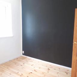 BK Wilbud Sp. z o.o. - Pokój po odświeżeniu: jedna ściana pomalowana na ciemny grafit, jasna drewniana podłoga, białe ściany z oknem i drewniane drzwi z przeszkleniem.