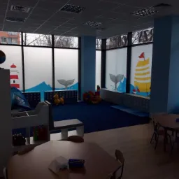 Wnętrze pokoju dziecięcego z motywem morskim: okna z grafikami latarni morskiej, ogon&oacute;w wieloryb&oacute;w i żagl&oacute;wki, niebieskie ściany i dywan, drewniane stoliki i krzesła, zabawki.