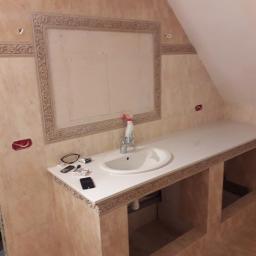 BK Wilbud Sp. z o.o. - Łazienka w trakcie wykańczania: beżowe płytki na ścianach, biała umywalka z baterią, biały blat z dekoracyjnym wykończeniem. Na blacie leżą okulary, papierosy i telefon. Widoczne niedokończone prace.