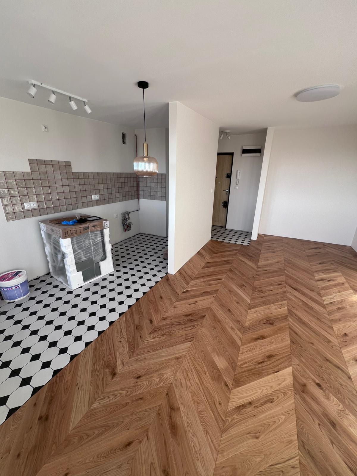 Wnętrze mieszkania w trakcie prac wykończeniowych: parkiet ułożony we wzór jodełki, fragment ściany z ułożonymi płytkami, zabezpieczona folią umywalka, wiaderko z farbą i czarno-biała podłoga w romby.