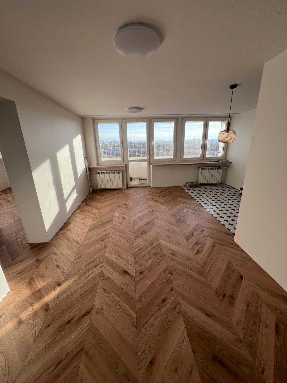 Jasne, przestronne wnętrze po remoncie z parkietem ułożonym w jodełkę, widok na miasto z okien i fragment wykończonej podłogi w czarno-białą szachownicę.