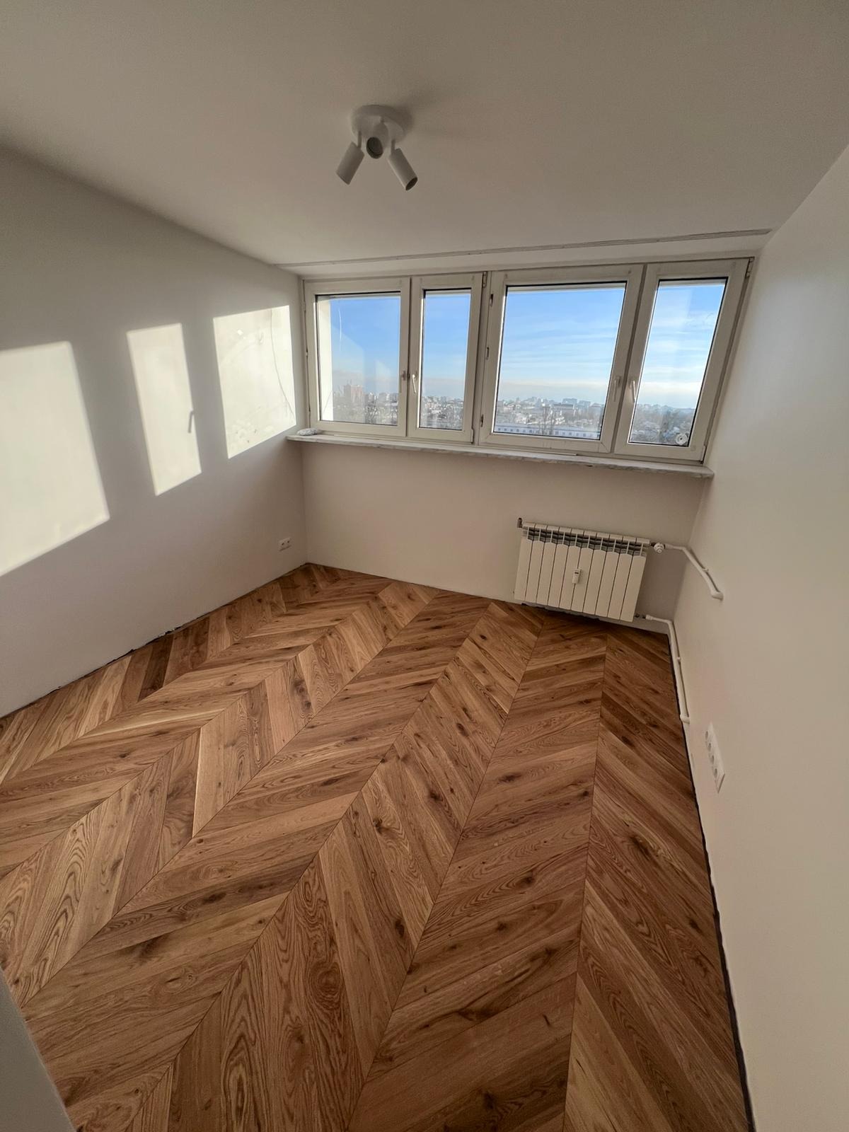 Pokój po remoncie z parkietem ułożonym we wzór jodełki, białe ściany, grzejnik pod oknem z widokiem na miasto.