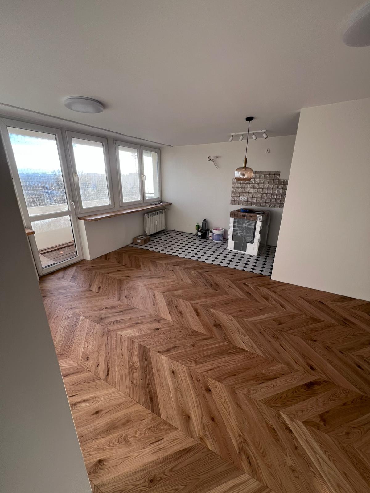 Nowoczesne mieszkanie w trakcie prac wykończeniowych: ułożona drewniana podłoga w jodełkę, widoczne okna balkonowe, grzejnik, fragment kuchni z płytkami w szachownicę oraz sprzęt AGD zabezpieczony...