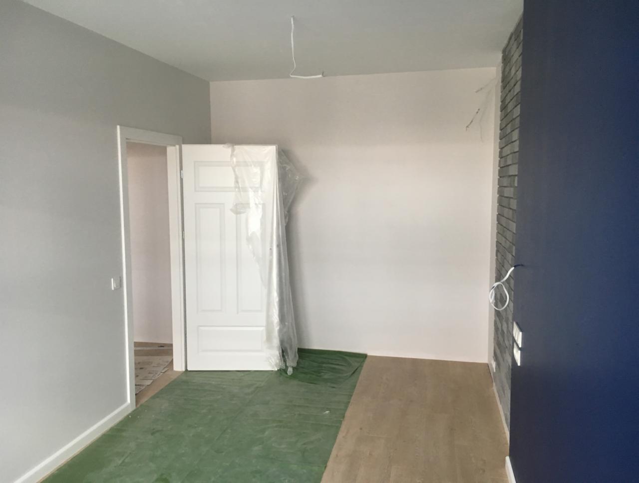 Wnętrze w trakcie prac wykończeniowych: zabezpieczona folią nowa biała ościeżnica, fragment ściany z imitacją cegły, przygotowane podłoże pod panele, szare i białe ściany.