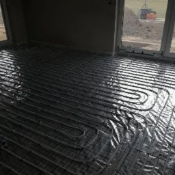 Układanie rur ogrzewania podłogowego na folii z siatką w surowym pomieszczeniu z oknami, widok na plac budowy za oknem.