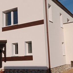 Usługi Ogólnobudowlane Dawid Niełacny - Świeżo otynkowana i pomalowana elewacja domu jednorodzinnego w kolorze jasnego beżu, z brązowymi elementami dekoracyjnymi i rynnami, widoczny pracownik na drabinie oraz leżący pies.