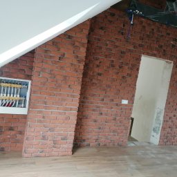 Usługi Ogólnobudowlane Dawid Niełacny - Ściana z cegły klinkierowej w pomieszczeniu na poddaszu, widoczna instalacja grzewcza w obudowie, fragment drewnianej podłogi i otwór drzwiowy.