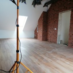 Usługi Ogólnobudowlane Dawid Niełacny - Poddasze po remoncie, wykończone cegłą dekoracyjną i panelami podłogowymi imitującymi drewno, oświetlone lampą budowlaną na statywie. Ślady po remoncie widoczne na podłodze.