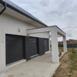 Nowoczesny dom z białymi ścianami, czarnym dachem i roletami zewnętrznymi, widok z boku, częściowo wykończona elewacja z widocznymi elementami ocieplenia.