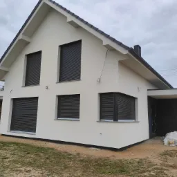 Nowoczesny dom jednorodzinny z jasną elewacją i antracytowymi roletami zewnętrznymi, widoczne worki i puszki po materiałach budowlanych wskazujące na prace wykończeniowe.