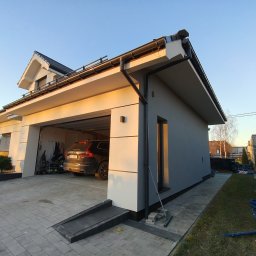 Usługi Ogólnobudowlane Dawid Niełacny - Nowoczesny dom z garażem wbudowanym, świeżo po malowaniu elewacji na szaro, z czarnymi detalami dachu i rynien. Widoczne kostki brukowe na podjeździe i fragment ogrodu.