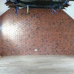 Usługi Ogólnobudowlane Dawid Niełacny - Ściana z cegły dekoracyjnej w poddaszu, z widocznym oświetleniem roboczym i zabezpieczeniem sufitu folią malarską, jasna podłoga imitująca drewno.
