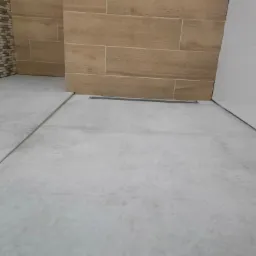 Wykończona łazienka z jasnymi płytkami podłogowymi imitującymi beton, ściana wyłożona płytkami imitującymi drewno oraz fragment ściany z mozaiką.