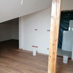 Usługi Ogólnobudowlane Dawid Niełacny - Poddasze po malowaniu ścian na biało, z widoczną drewnianą podłogą, fragmentem drewnianej belki, puszkami z farbą i przygotowanymi otworami na gniazdka elektryczne.