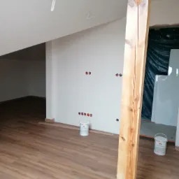 Poddasze po malowaniu ścian na biało, z widoczną drewnianą podłogą, fragmentem drewnianej belki, puszkami z farbą i przygotowanymi otworami na gniazdka elektryczne.