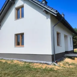 Świeżo pomalowana, biała elewacja domu jednorodzinnego z szarym cokołem, brązowe okna i szara dachówka, widok z zewnątrz w słoneczny dzień.