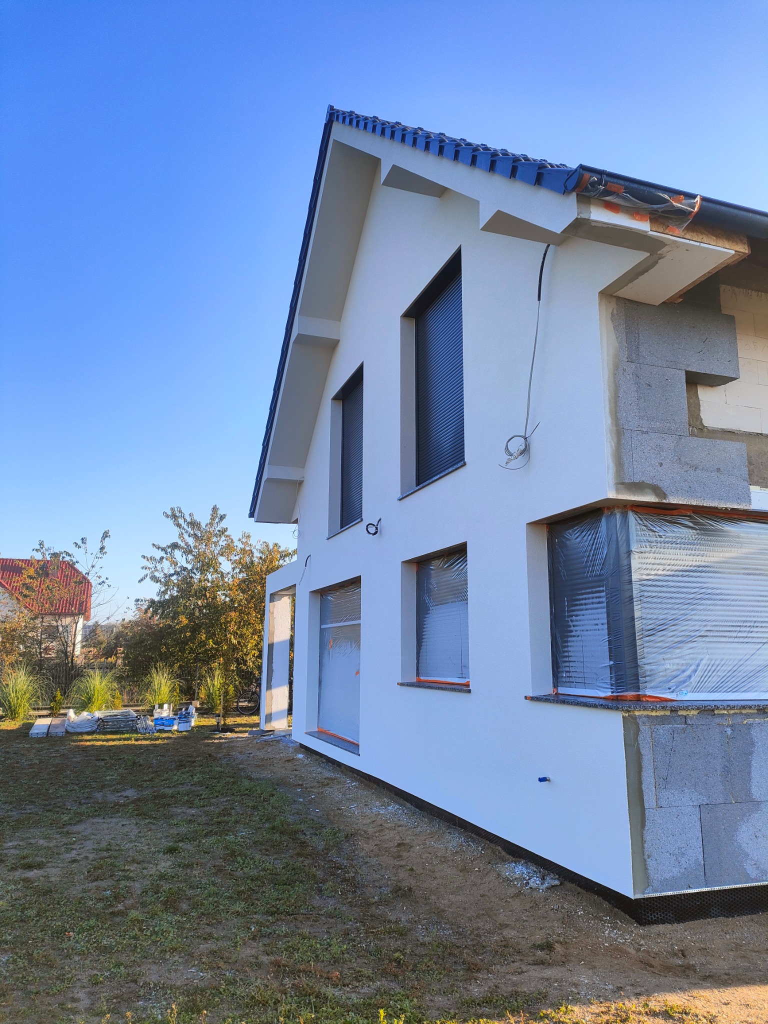 Niedokończona elewacja domu jednorodzinnego z widocznym ociepleniem w narożniku, okna zabezpieczone folią, tynk w kolorze białym, fragment dachu z czarną dachówką, trawnik przed budynkiem.