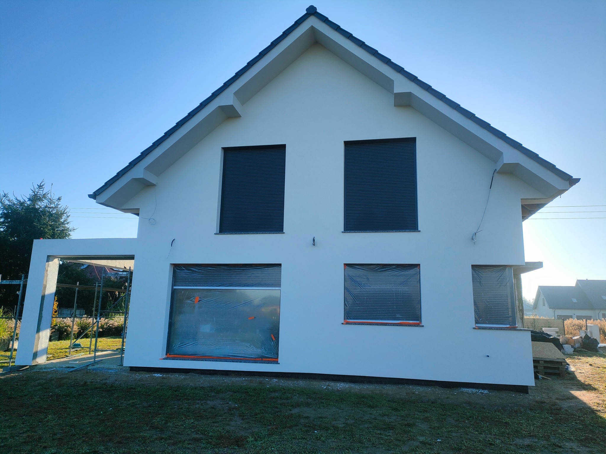 Nowoczesny dom jednorodzinny z białą elewacją, okna zabezpieczone folią malarską, widoczne rusztowanie i materiały budowlane, błękitne niebo w tle.