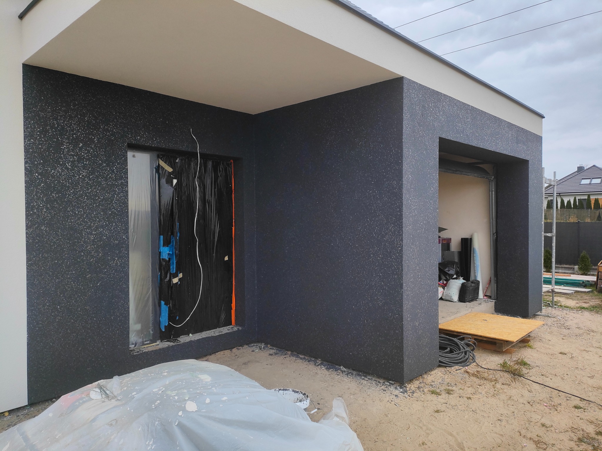 Nowoczesny budynek z elewacją w odcieniach szarości, okno zabezpieczone folią malarską, widoczne przygotowania do prac wykończeniowych na zewnątrz.