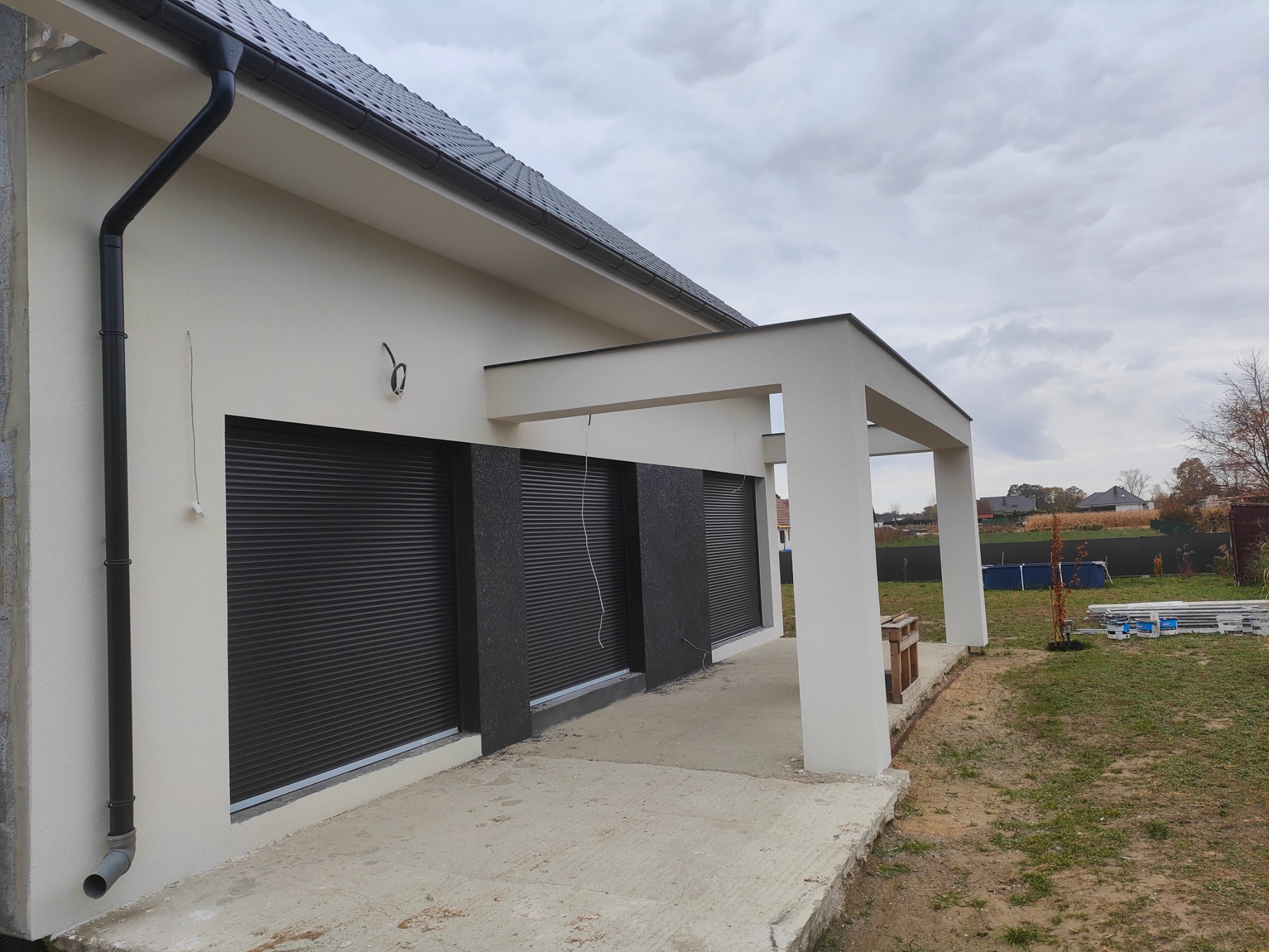 Nowoczesny dom z białymi ścianami, czarnym dachem i roletami zewnętrznymi, widok z boku, częściowo wykończona elewacja z widocznymi elementami ocieplenia.