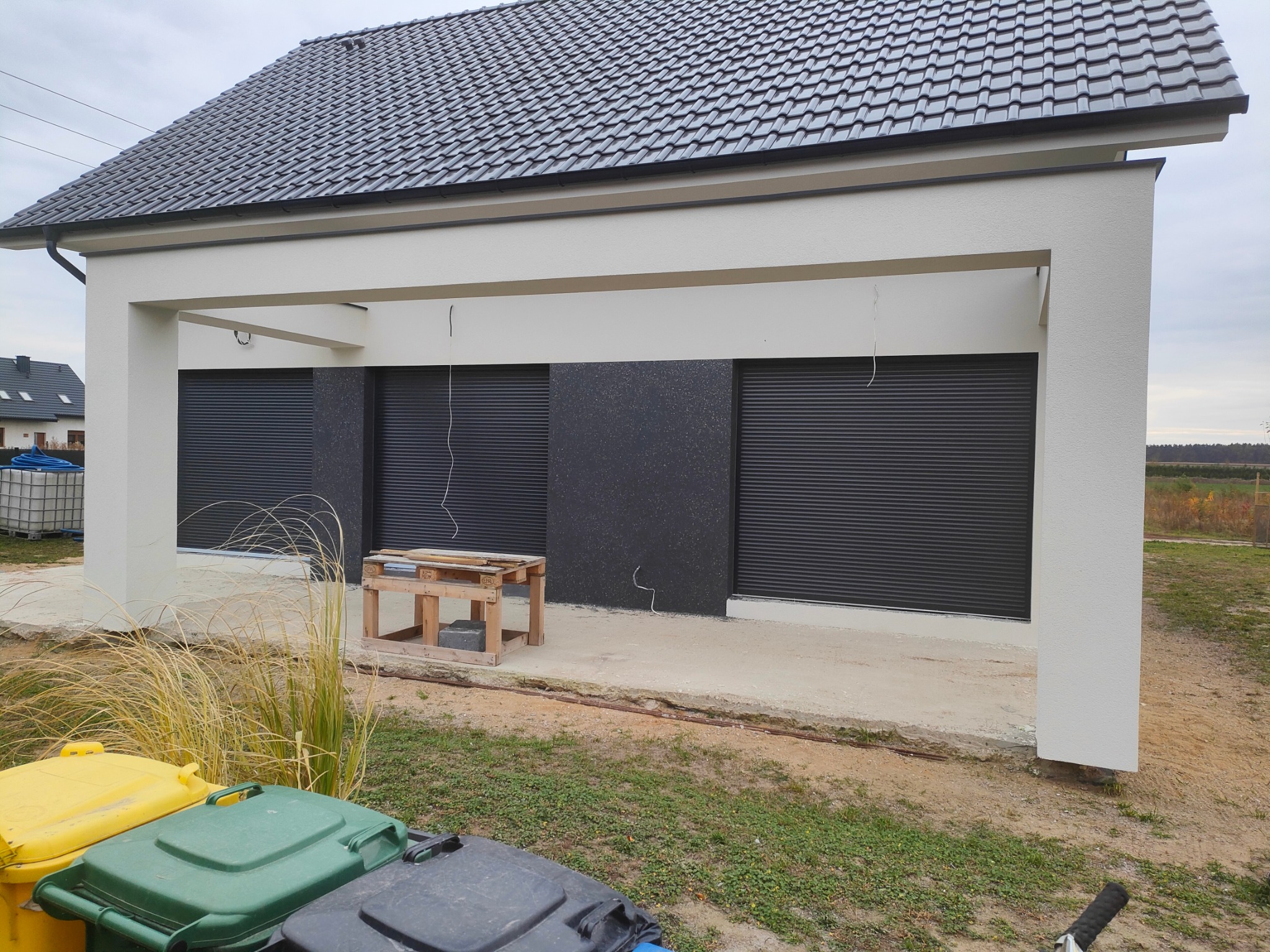 Nowoczesny dom z białymi ścianami i grafitowymi roletami zewnętrznymi; na pierwszym planie widoczne kosze na śmieci.