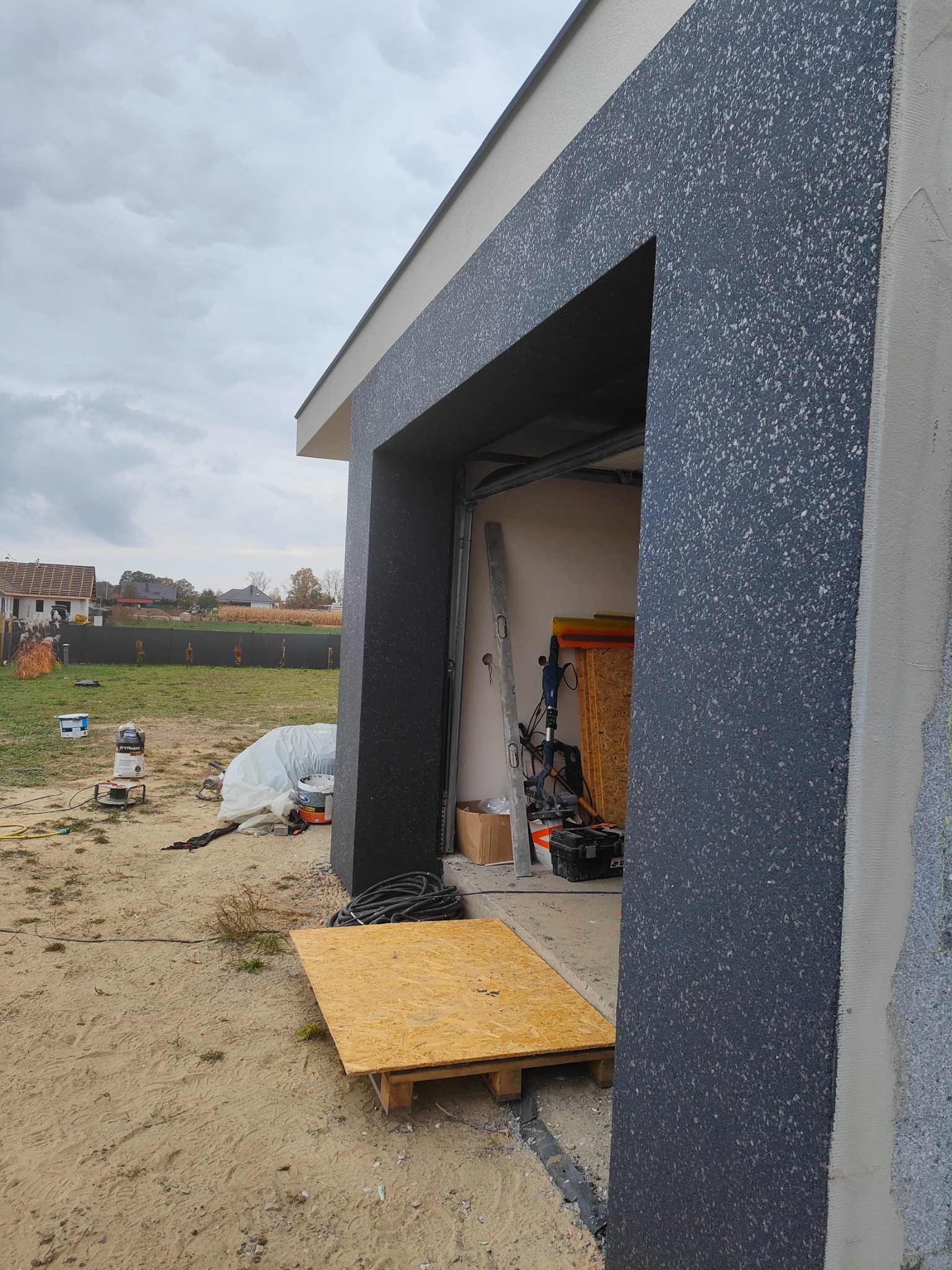 Elewacja budynku z wykończonym fragmentem w kolorze grafitowym z fakturą, w tle widoczne narzędzia i materiały budowlane, częściowo ocieplona ściana, paleta z płytą OSB na piasku.