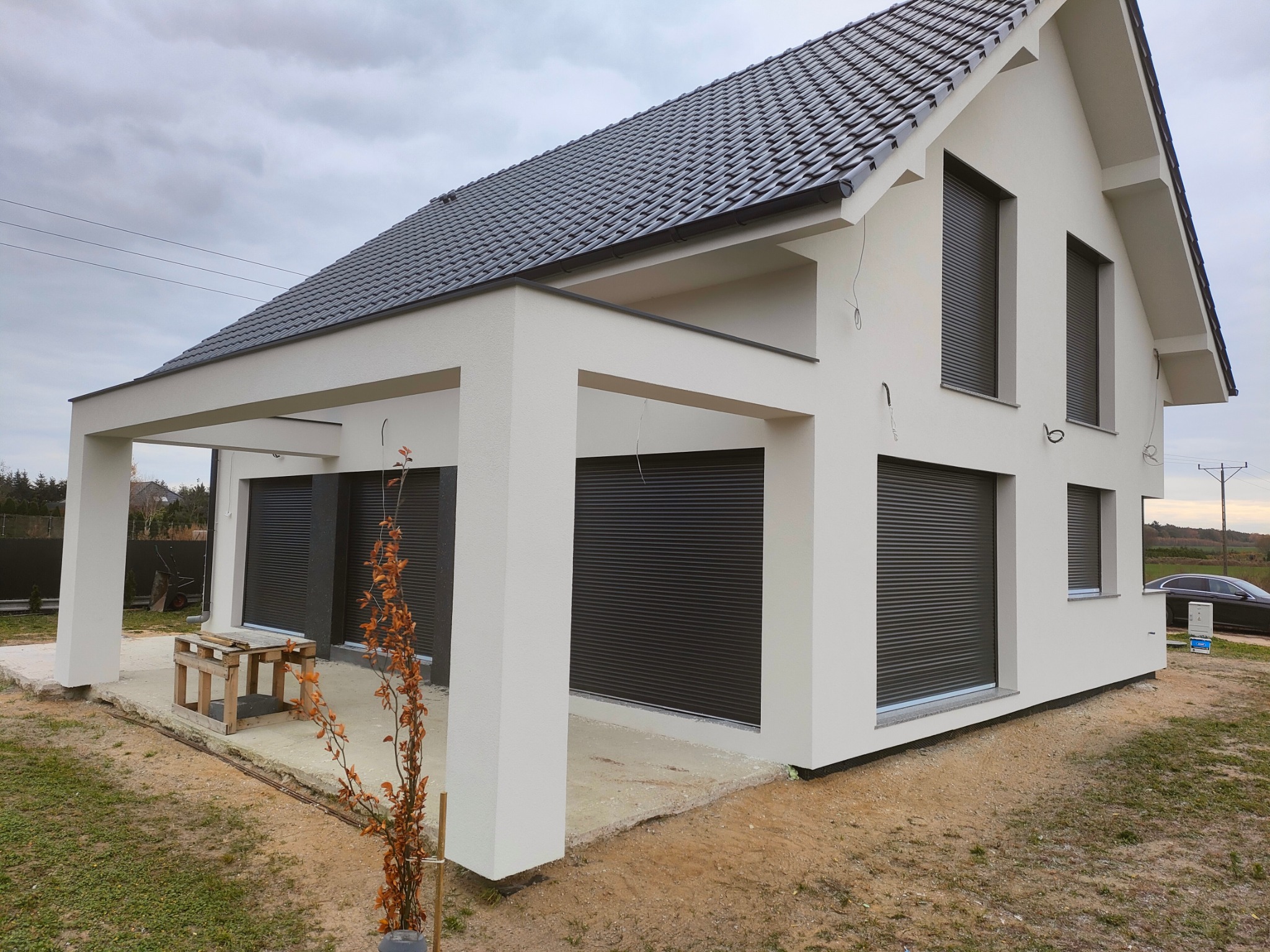 Nowoczesny dom jednorodzinny z białymi ścianami i grafitową dachówką oraz roletami zewnętrznymi, widok z perspektywy ogrodu, z widocznym fragmentem niedokończonego chodnika i drewnianym stołem...