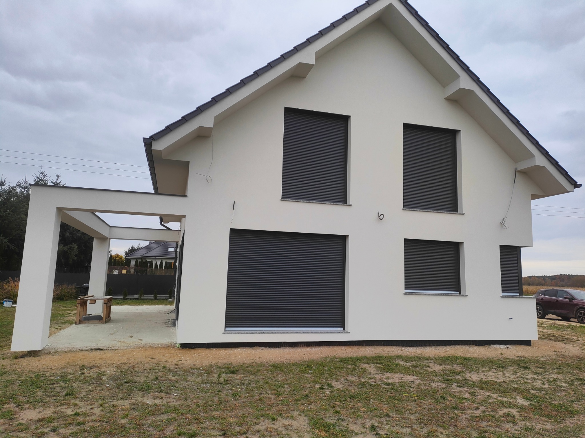 Nowoczesny dom jednorodzinny z białym tynkiem elewacyjnym i szarymi roletami zewnętrznymi, widok z zewnątrz na świeżo wykończoną elewację i częściowo zagospodarowany teren.