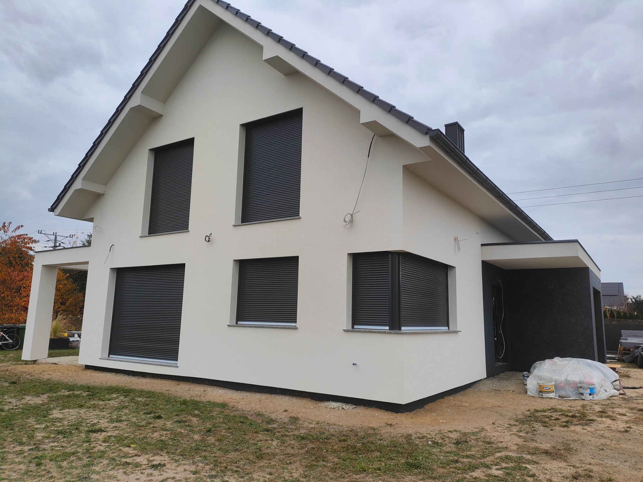 Nowoczesny dom jednorodzinny z jasną elewacją i antracytowymi roletami zewnętrznymi, widoczne worki i puszki po materiałach budowlanych wskazujące na prace wykończeniowe.