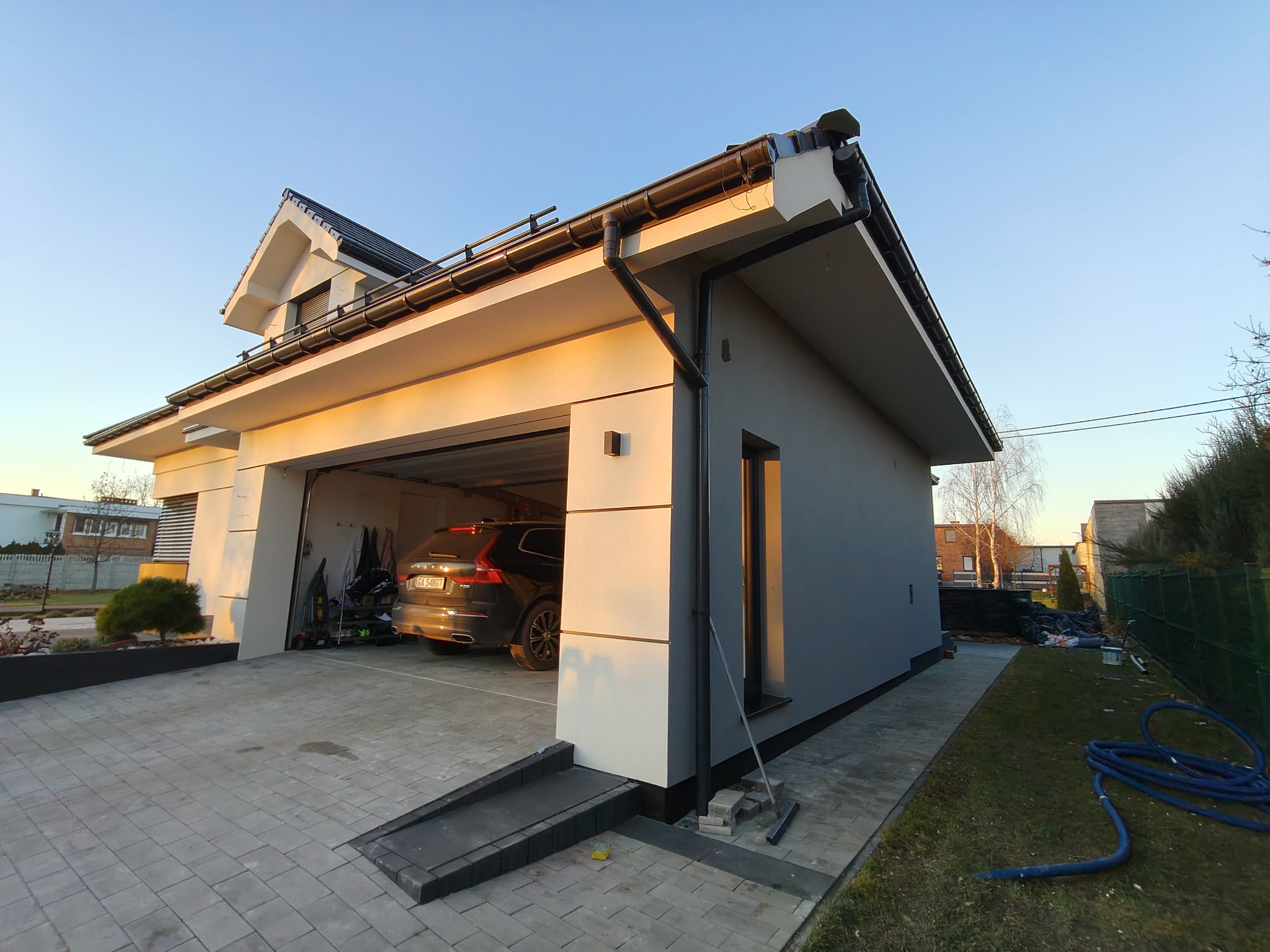 Nowoczesny dom z garażem wbudowanym, świeżo po malowaniu elewacji na szaro, z czarnymi detalami dachu i rynien. Widoczne kostki brukowe na podjeździe i fragment ogrodu.