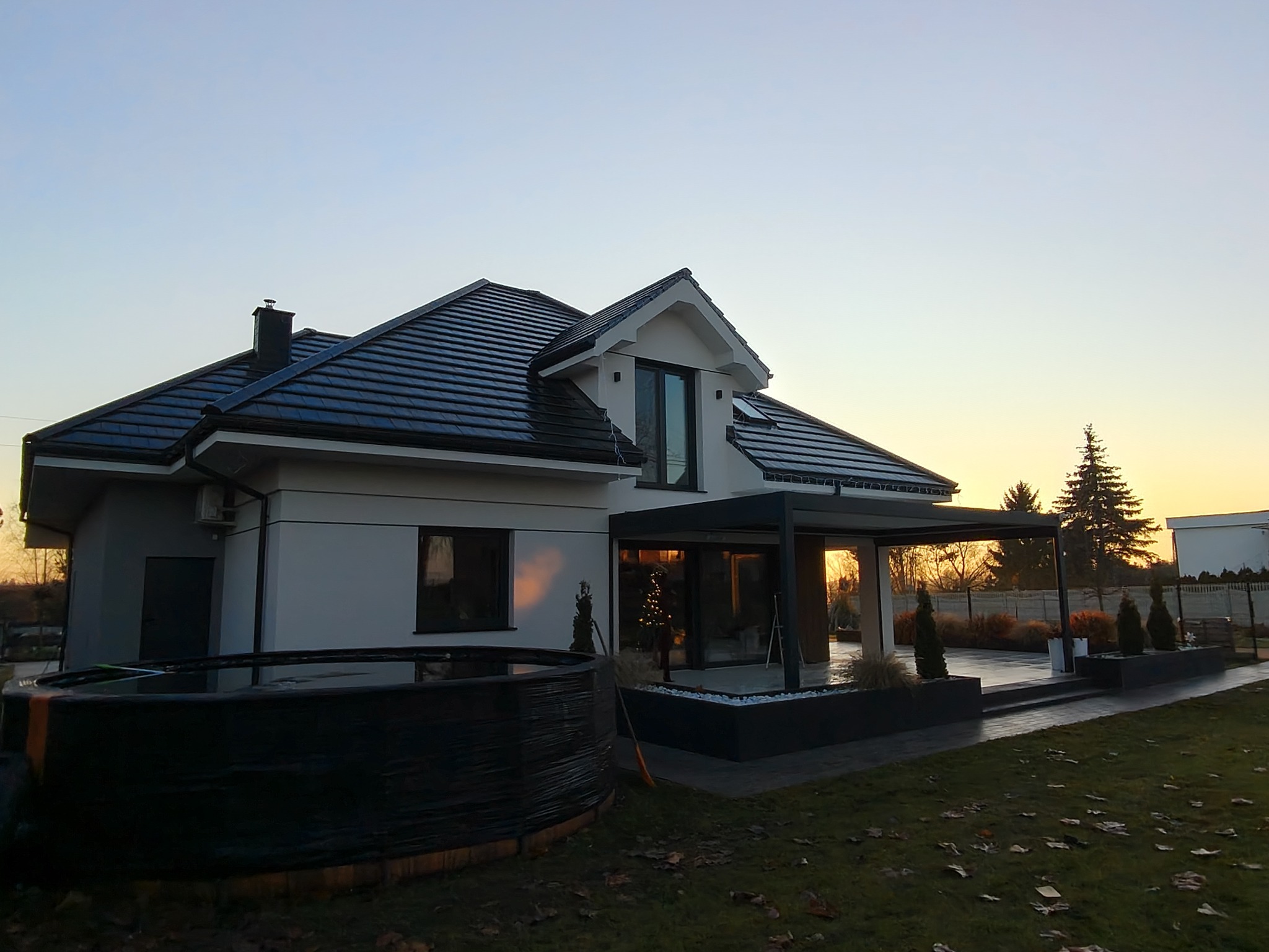 Nowoczesny dom jednorodzinny z białymi ścianami i ciemną dachówką, widoczny z ogrodu o zmierzchu. Na pierwszym planie basen ogrodowy.