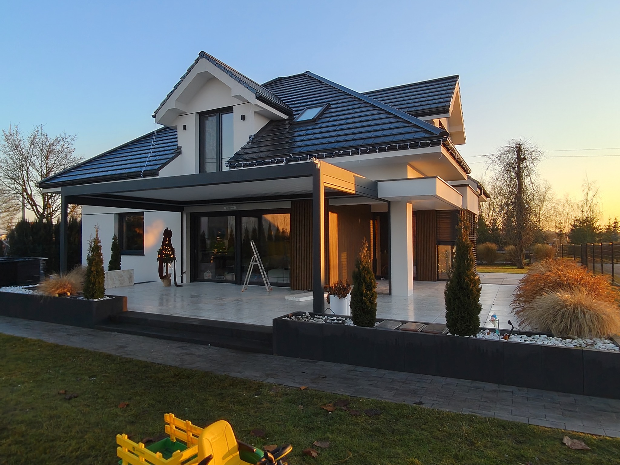 Nowoczesny dom jednorodzinny z pergolą i tarasem w ciepłym świetle zachodzącego słońca. Widoczne elementy wykończenia elewacji i zagospodarowania terenu.