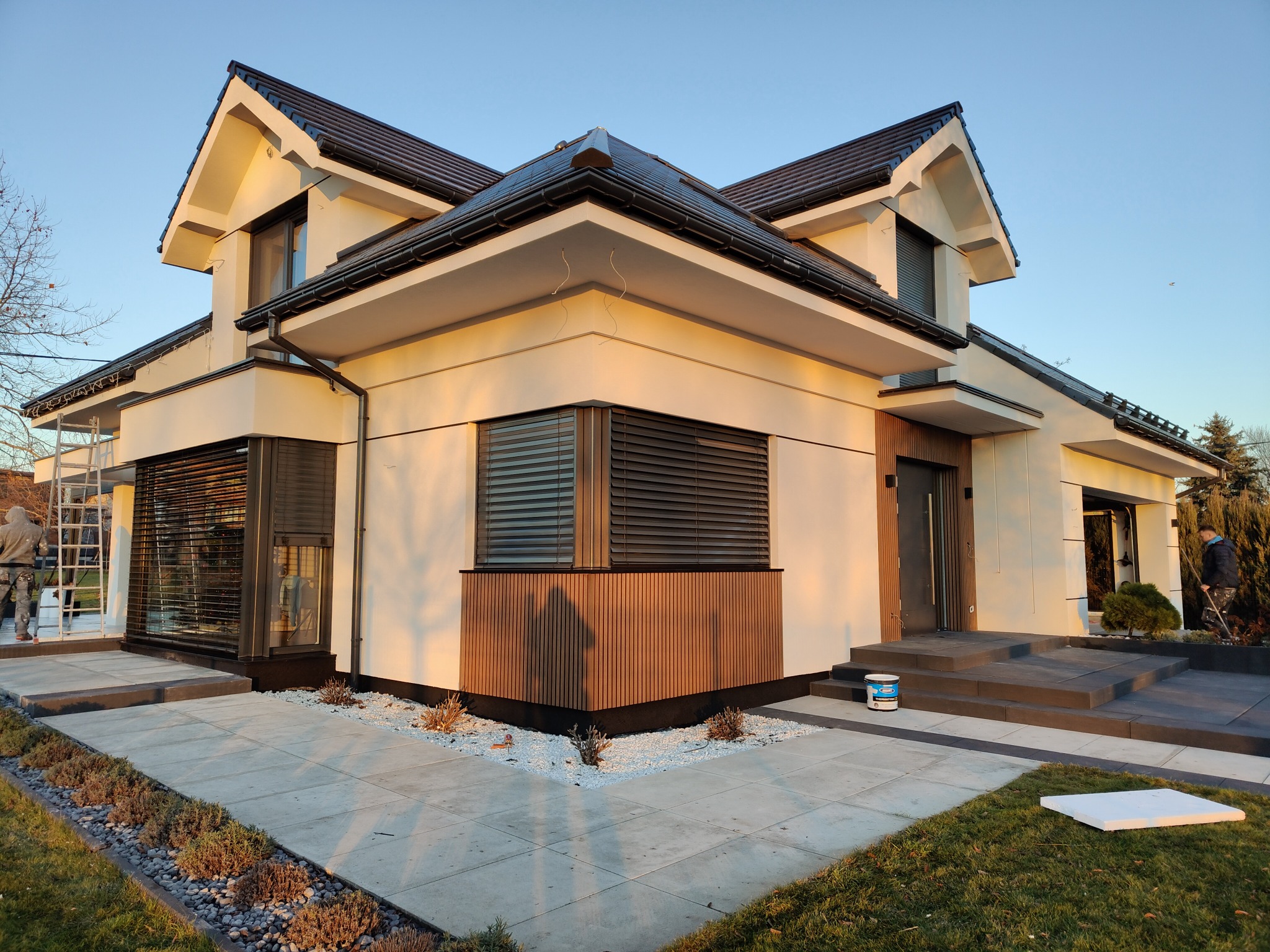 Nowoczesny dom jednorodzinny z jasną elewacją i ciemną stolarką okienną, widok z zewnątrz, z elementami wykończenia elewacji i zagospodarowania terenu.