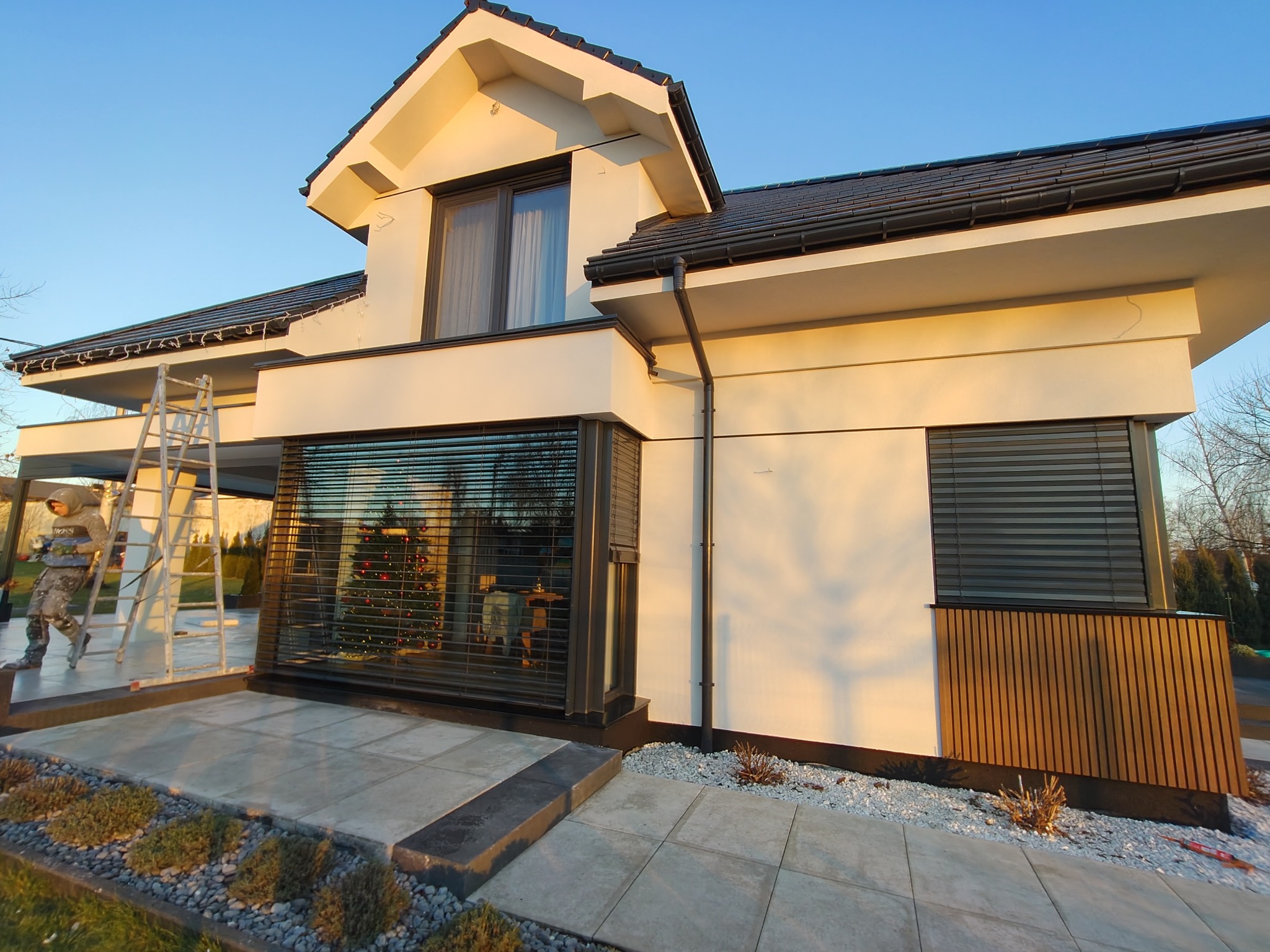 Nowoczesny dom jednorodzinny z białymi ścianami i elementami z drewna. Widoczna drabina i pracownik. W oknach żaluzje zewnętrzne. Świąteczne dekoracje wewnątrz.