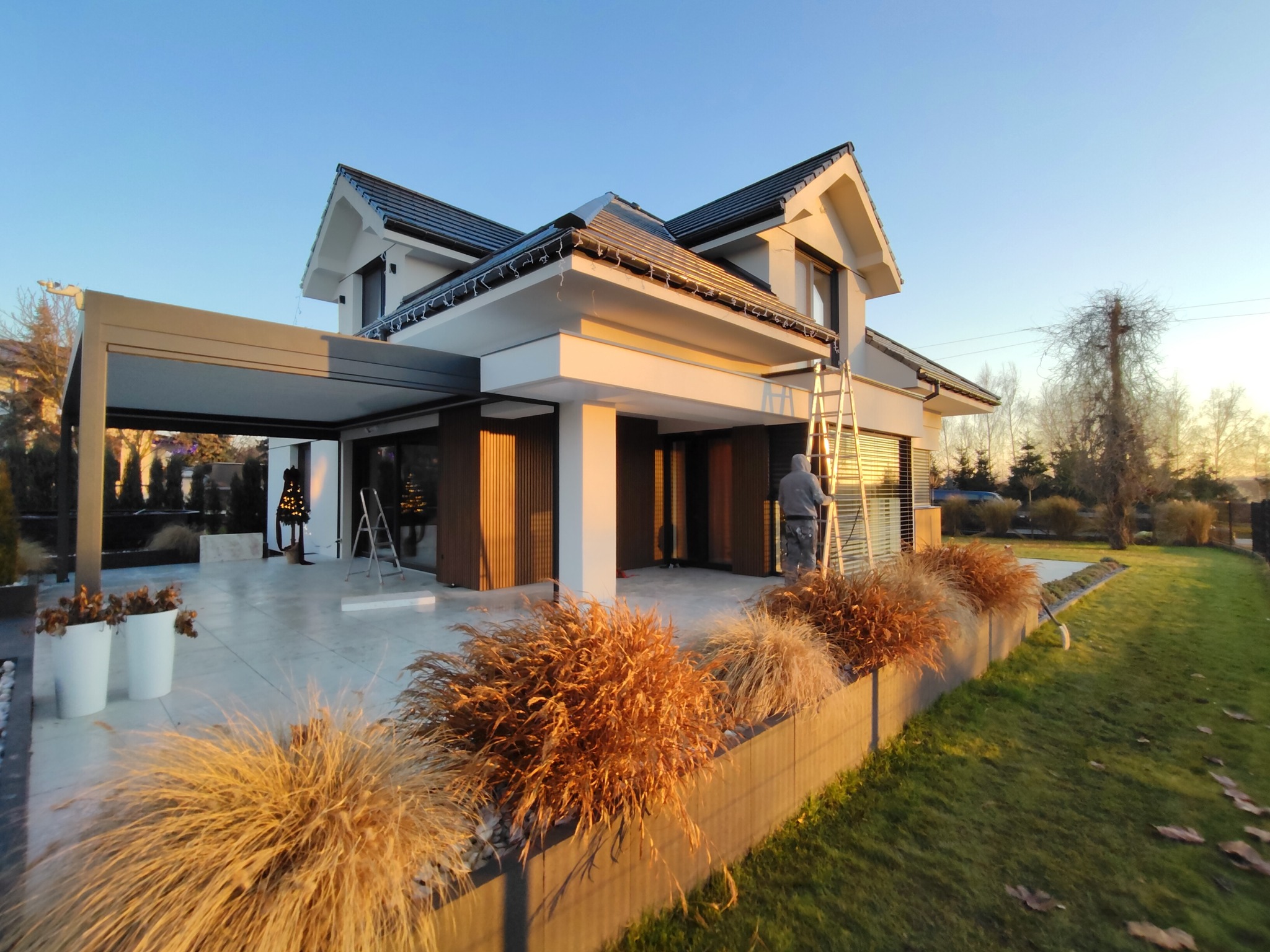 Nowoczesny dom jednorodzinny z pergolą i elementami drewnianymi, oświetlony ciepłym światłem zachodzącego słońca. Pracownik na drabinie przy oknie. Dekoracyjne trawy wzdłuż ogrodzenia.
