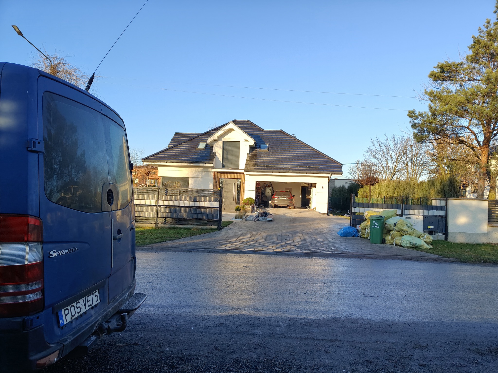 Dom jednorodzinny z podjazdem wyłożonym kostką, widoczny samochód dostawczy i worki z odpadami. Prace wykończeniowe w toku, słoneczny dzień.