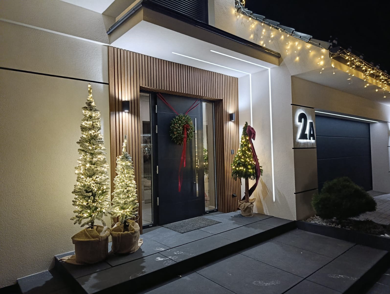 Elegancka elewacja domu z drewnianą okładziną, oświetleniem LED i świątecznymi dekoracjami: wieniec na drzwiach, choinki z lampkami. Nowoczesny design.